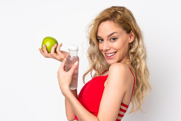 Jeune femme blonde isolée sur un mur blanc avec une pomme et avec une bouteille d'eau