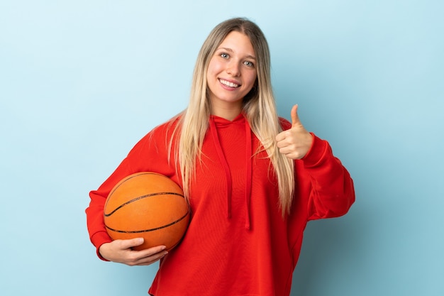 Jeune femme blonde isolée sur mur bleu jouant au basket et avec le pouce vers le haut