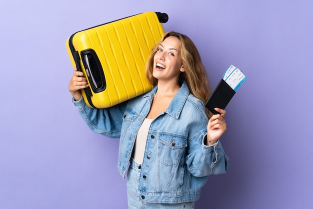 Jeune femme blonde isolée sur un mur violet en vacances avec valise et passeport