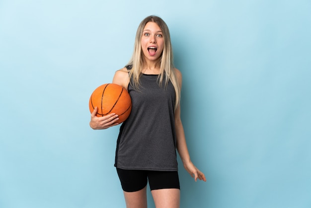 Jeune femme blonde jouant au basket-ball isolé sur mur bleu avec une expression faciale surprise