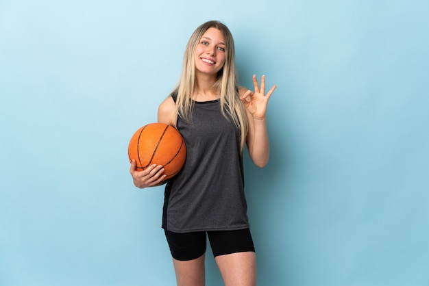 Jeune femme blonde jouant au basketball isolé sur mur bleu montrant un signe ok avec les doigts