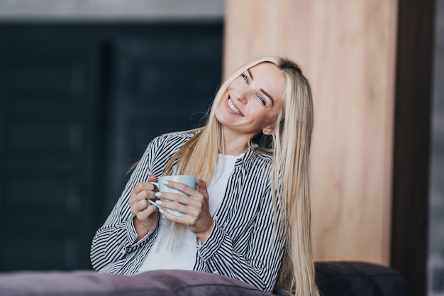 Une jeune femme blonde joyeuse en tenue décontractée tient une tasse de café de larges sourires regarde la caméra heureusement assise sur un canapé confortable à la maison Une magnifique étudiante suédoise profite du week-end au matin ensoleillé Belles personnes