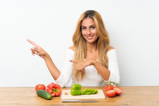 Jeune femme blonde avec des légumes dans une table, pointant le doigt sur le côté