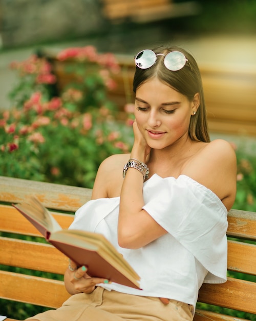 Jeune femme blonde lisant un livre avec enthousiasme alors qu'il était assis sur un banc dans le parc