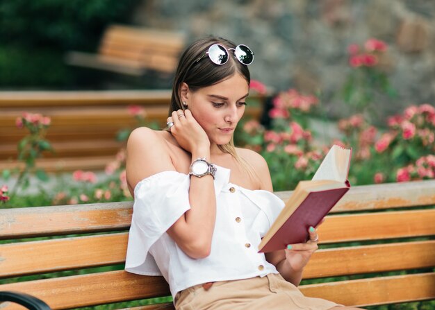 Jeune femme blonde lisant un livre avec enthousiasme alors qu'il était assis sur un banc dans le parc