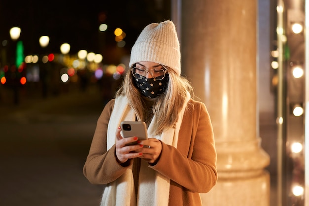 Jeune femme blonde avec un masque devant une vitrine d'écrire un message, dans une ville la nuit. Ambiance hivernale.