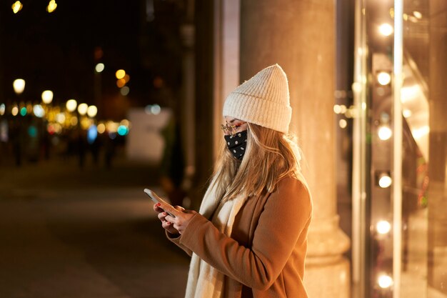 Jeune femme blonde avec un masque devant une vitrine de magasin d'écrire un message, dans une ville la nuit, avec des lumières en arrière-plan. Ambiance hivernale.