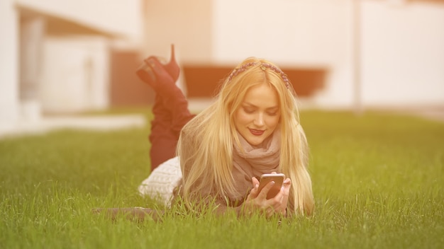 Jeune femme blonde mignonne élégante avec du maquillage se trouve dans la pelouse verte et regarde le smartphone sur fond de bâtiment flou, lumière du soleil.