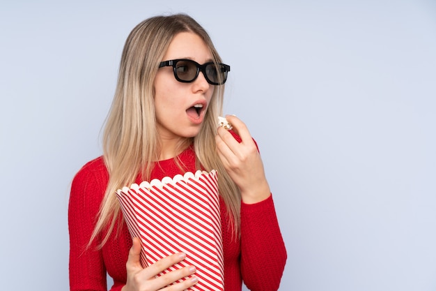 Jeune femme blonde sur un mur bleu isolé avec des lunettes 3d et tenant un grand seau de pop-corn tout en regardant de côté