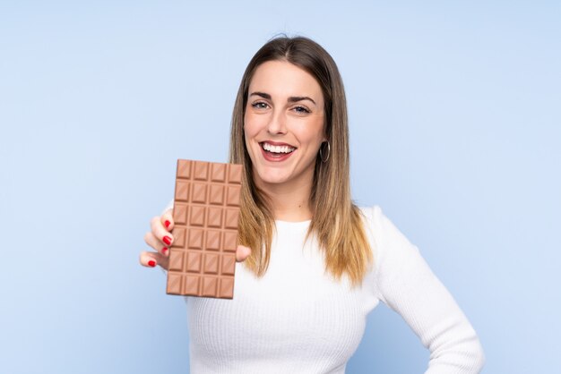 Jeune femme blonde sur un mur bleu isolé en prenant une tablette de chocolat et heureux