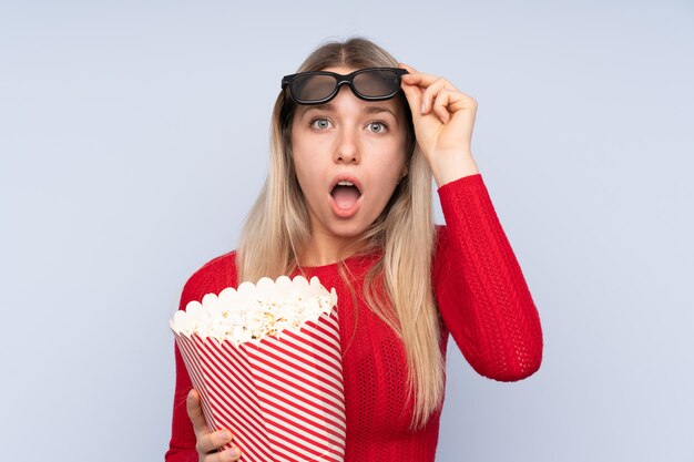 Jeune femme blonde sur mur bleu isolé surpris avec des lunettes 3d et tenant un grand seau de pop-corn