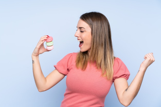 Jeune femme blonde sur un mur bleu isolé tenant des macarons français colorés et célébrant une victoire