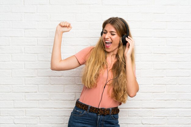 Jeune Femme Blonde Sur Le Mur De Briques Blanches, écouter De La Musique Avec Des écouteurs