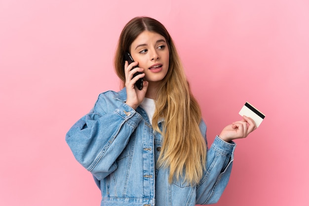 Jeune femme blonde sur un mur isolé en gardant une conversation avec le téléphone mobile et tenant une carte de crédit