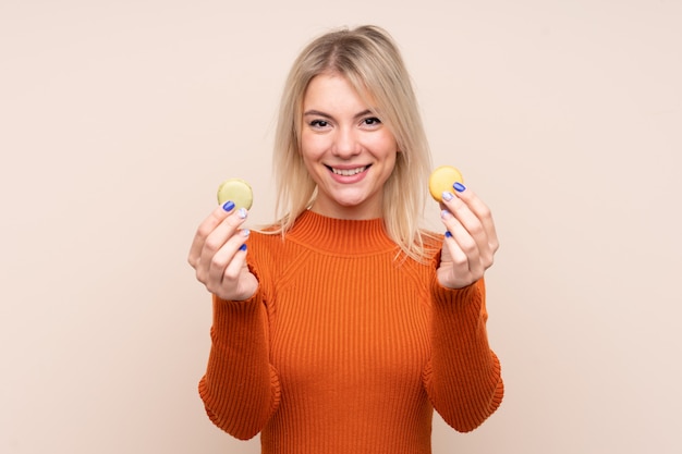 Jeune femme blonde sur un mur isolé offrant des macarons français colorés