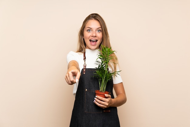 Jeune femme blonde sur un mur isolé prenant un pot de fleurs et pointant vers l'avant