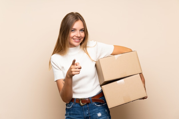 Jeune femme blonde sur un mur isolé tenant une boîte pour la déplacer sur un autre site tout en pointant vers l'avant