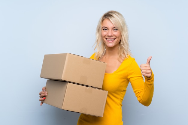 Jeune femme blonde sur mur isolé tenant une boîte pour le déplacer vers un autre site avec le pouce vers le haut