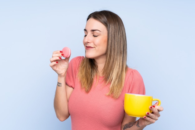 Jeune femme blonde sur un mur isolé tenant des macarons français colorés et une tasse de lait