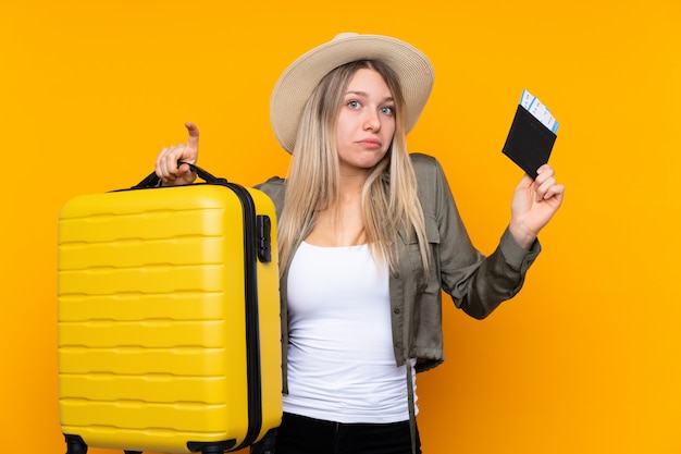 Jeune femme blonde sur mur jaune isolé malheureux en vacances avec valise et passeport