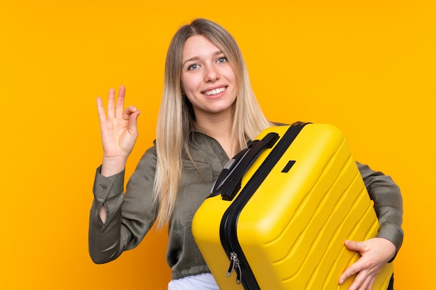 Jeune femme blonde sur mur jaune isolé en vacances avec valise de voyage et faisant signe OK