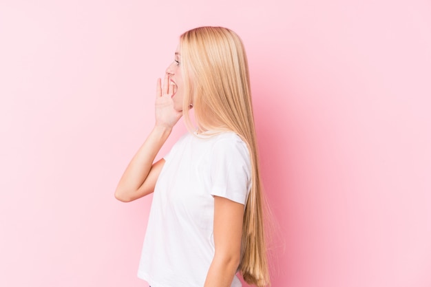 Jeune femme blonde sur mur rose criant et tenant la paume près de la bouche ouverte.