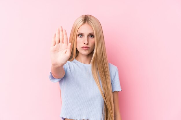 Jeune femme blonde sur mur rose debout avec la main tendue montrant le panneau d'arrêt, vous empêchant.