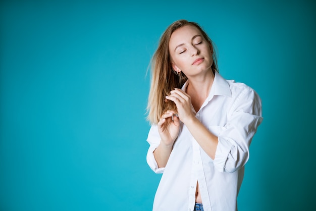 Jeune femme blonde d'origine ethnique caucasienne posant dans une chemise blanche et un jean sur un mur bleu