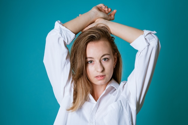 Jeune femme blonde d'origine ethnique caucasienne posant dans une chemise blanche et un jean sur un mur bleu