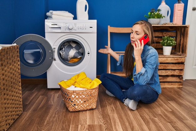 Jeune femme blonde parlant sur smartphone pour machine à laver cassée dans la buanderie