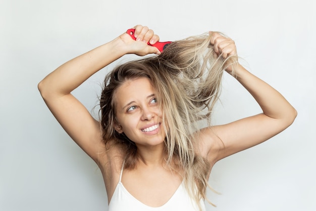 Une jeune femme blonde peignant ses cheveux décolorés secs indisciplinés emmêlés avec un peigne rouge