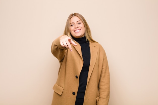 Jeune femme blonde pointant sur la caméra avec un sourire satisfait, confiant et amical, vous choisissant contre un mur plat