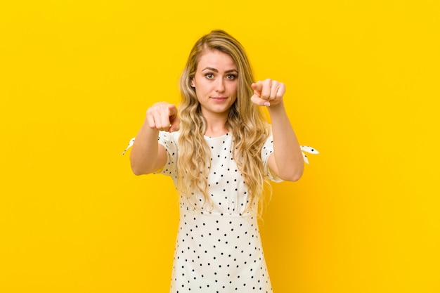 Jeune femme blonde pointant vers l'avant à la caméra avec les deux doigts et l'expression en colère, vous disant de faire votre devoir contre le mur jaune