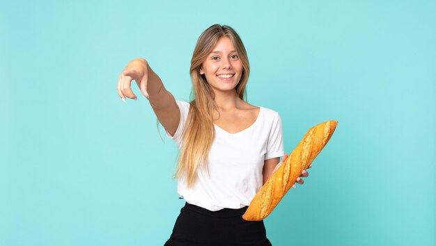 Jeune femme blonde pointant vers la caméra vous choisissant et tenant une baguette de pain