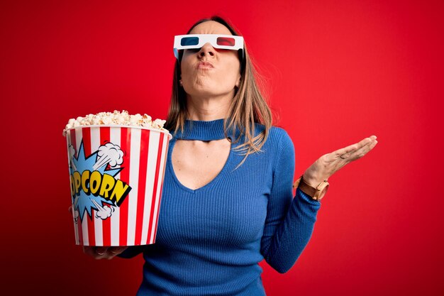 Jeune femme blonde portant des lunettes 3d et mangeant un paquet de pop-corn en regardant un film au cinéma fou et fou criant et hurlant avec une expression agressive et les bras levés Concept de frustration