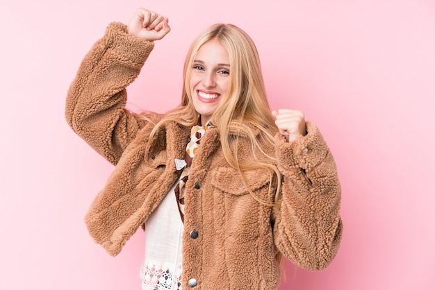 Jeune femme blonde portant un manteau contre un mur rose célébrant une journée spéciale, saute et lève les bras avec énergie.