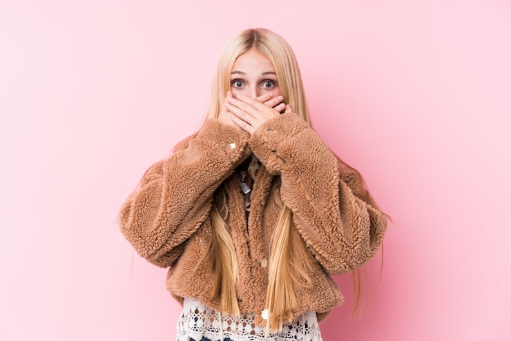 Photo jeune femme blonde portant un manteau contre un mur rose choqué couvrant la bouche avec les mains.
