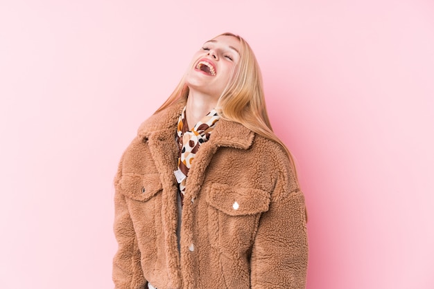 Jeune femme blonde portant un manteau contre un mur rose détendu et heureux de rire, le cou tendu montrant les dents.