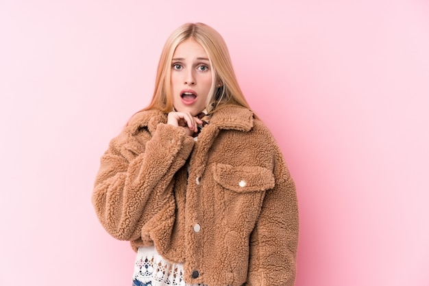 Jeune femme blonde portant un manteau contre un mur rose effrayé et effrayé.