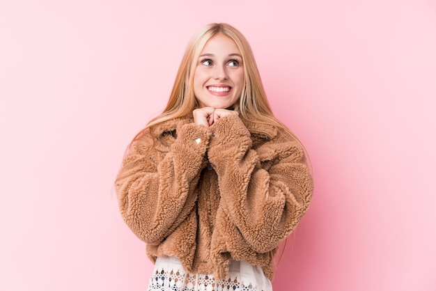 Une jeune femme blonde portant un manteau contre un mur rose garde les mains sous le menton, regarde joyeusement de côté.