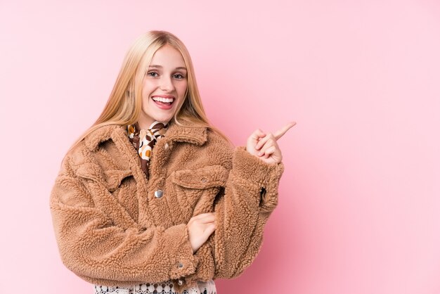 Jeune femme blonde portant un manteau sur un fond rose souriant joyeusement pointant avec l'index loin.