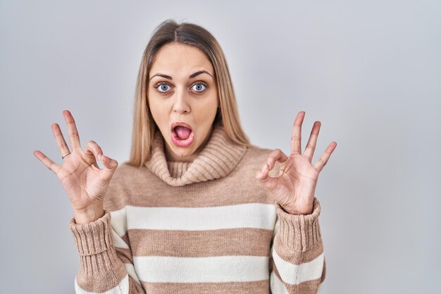 Jeune femme blonde portant un pull à col roulé sur fond isolé à la surprise et choquée de faire le symbole d'approbation ok avec les doigts. expression folle