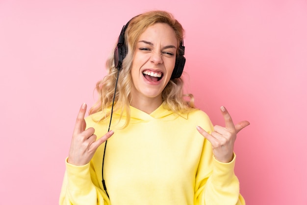 Jeune femme blonde portant un sweat-shirt isolé sur le mur rose en écoutant de la musique faisant un geste rock