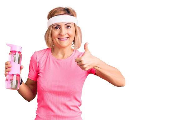 Jeune femme blonde portant des vêtements de sport tenant une bouteille d'eau souriant heureux et positif pouce vers le haut faisant un excellent signe d'approbation