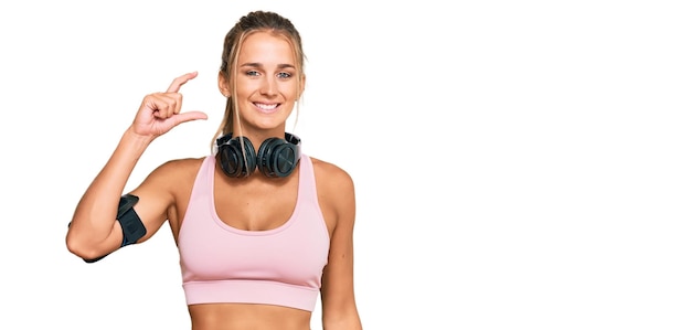 Jeune femme blonde portant des vêtements de sport et utilisant des écouteurs souriant et confiant gesticulant avec la main faisant un signe de petite taille avec les doigts regardant et le concept de mesure de la caméra