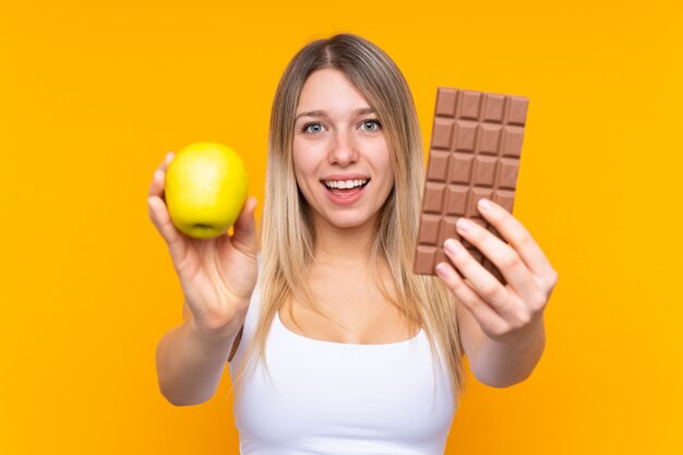 Jeune femme blonde prenant une tablette de chocolat dans une main et une pomme dans l'autre