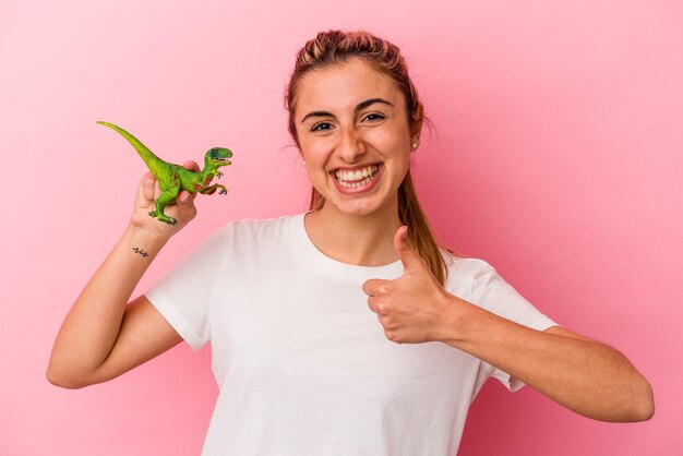 Jeune femme blonde de race blanche tenant une miniature de dinosaure isolée sur un mur rose en souriant et en levant le pouce vers le haut