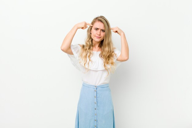 Jeune femme blonde avec un regard sérieux et concentré, remue-méninges et penser à un problème difficile contre le mur blanc