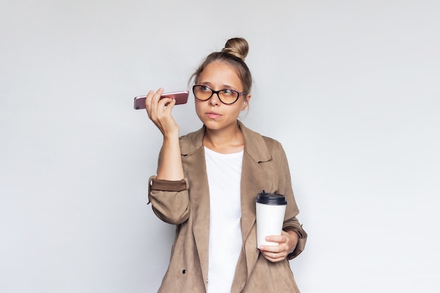 Une jeune femme blonde réussie tient une tasse de café ou de thé en papier blanc écoutant des messages vocaux