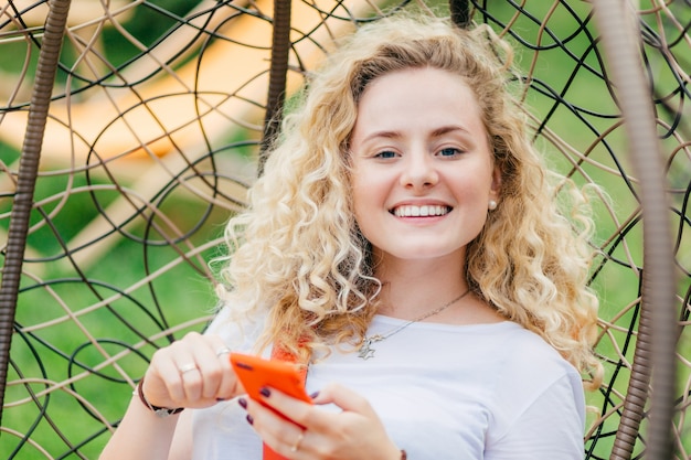 Jeune femme blonde satisfaite aux cheveux bouclés, a un sourire positif, utilise un téléphone portable moderne pour discuter avec des amis